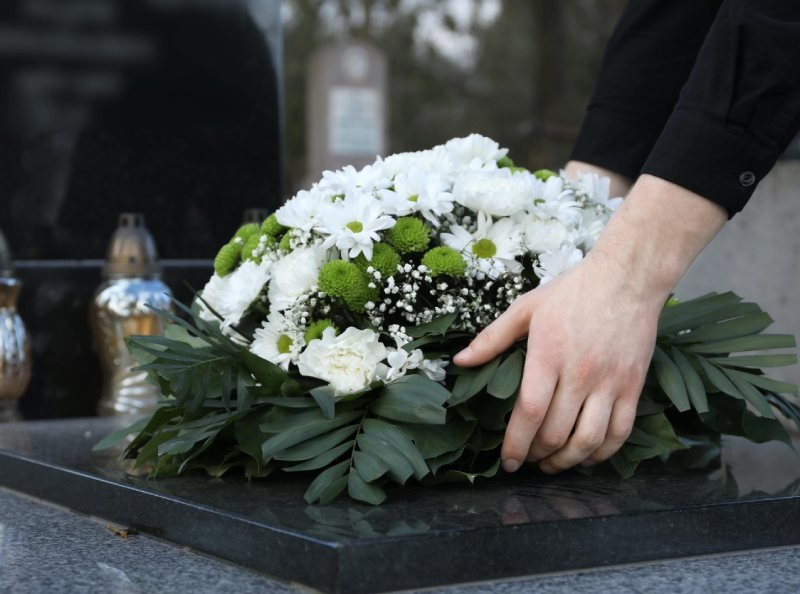 Woman putting funeral wreath of flowers on granite tombstone in cemetery closeup